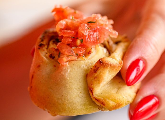 יד של אישה עם לק אדום מחזיקה רוזלך - גלגול של בצק במילוי גבינות ומעל רסק עגבניות, צילום: גליה אבירם